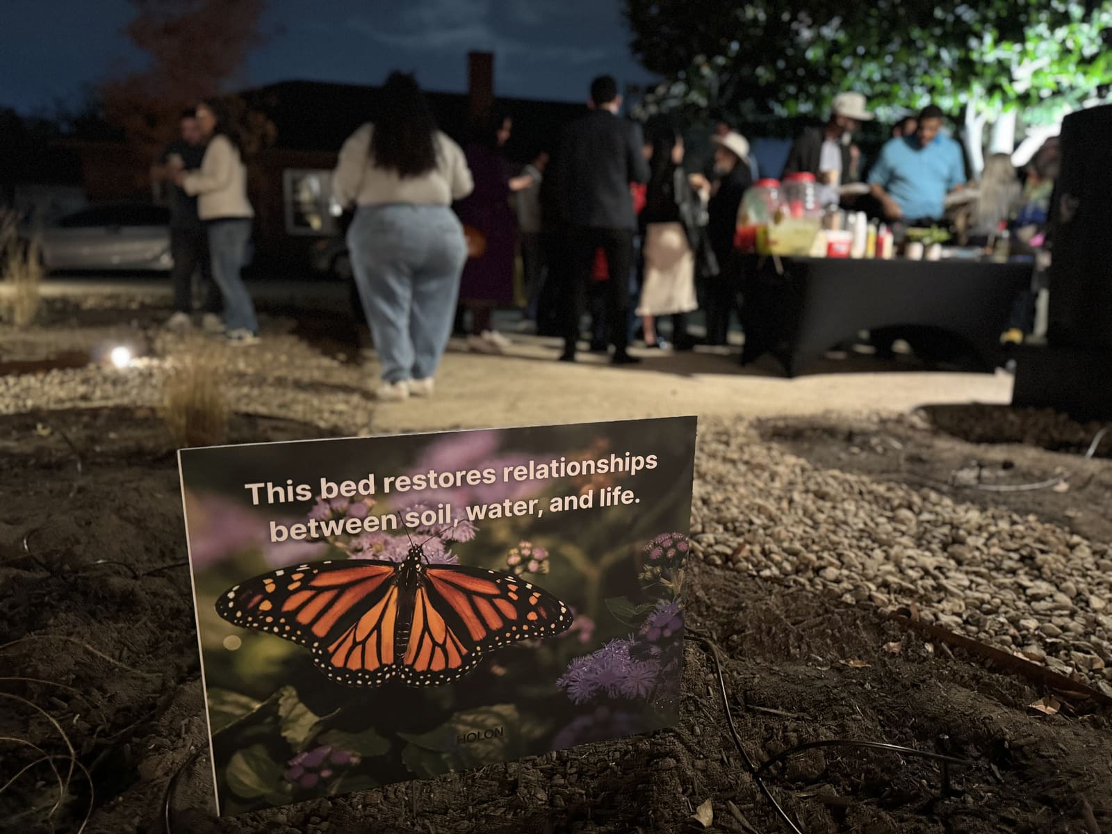 A garden sign with a monarch butterfly reading this bed restores relationships between soil, water, and life, people gathered in the evening behind