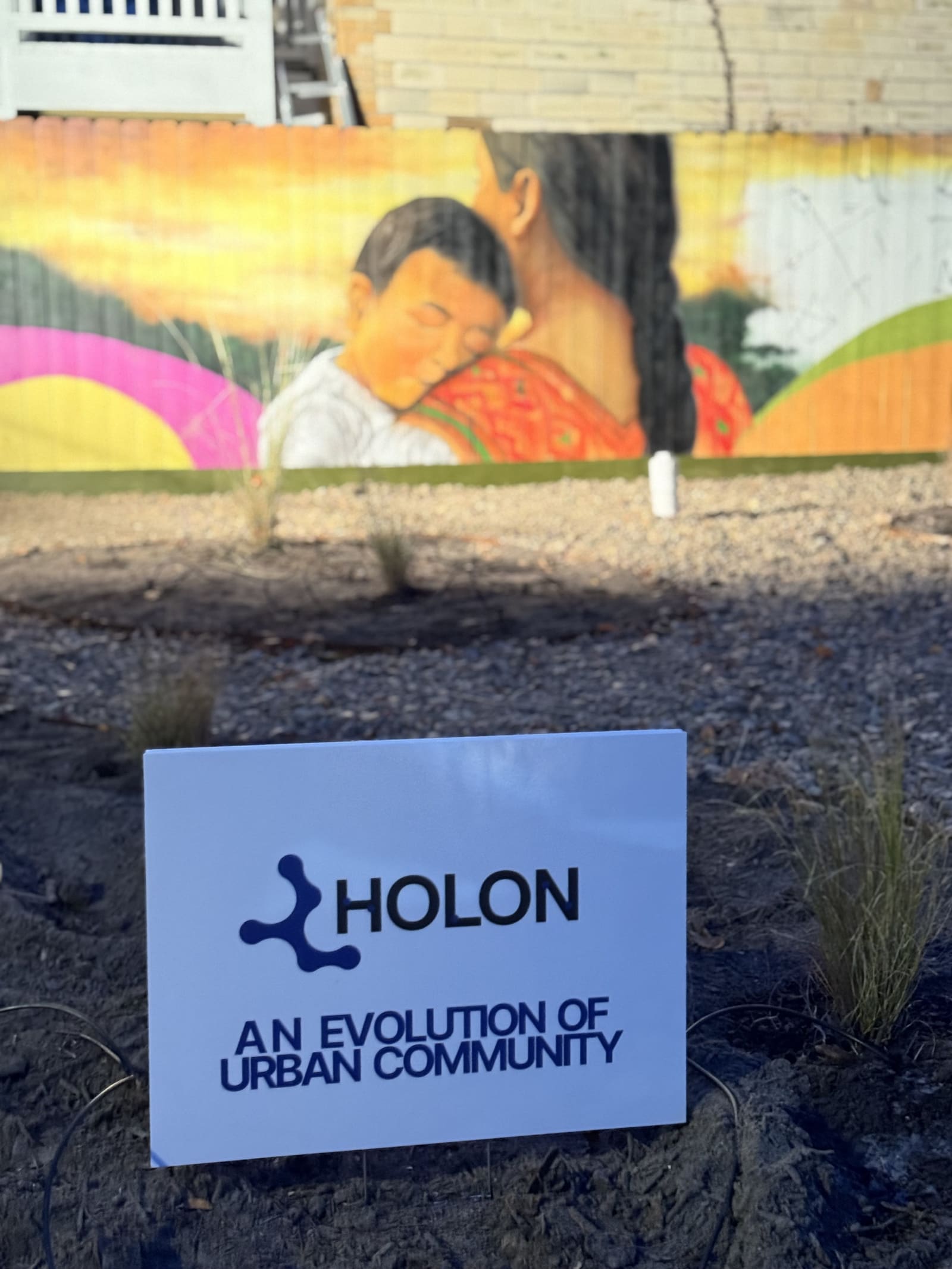 A small Holon sign staked in fresh mulch, a mural of a mother holding a child soft in the background
