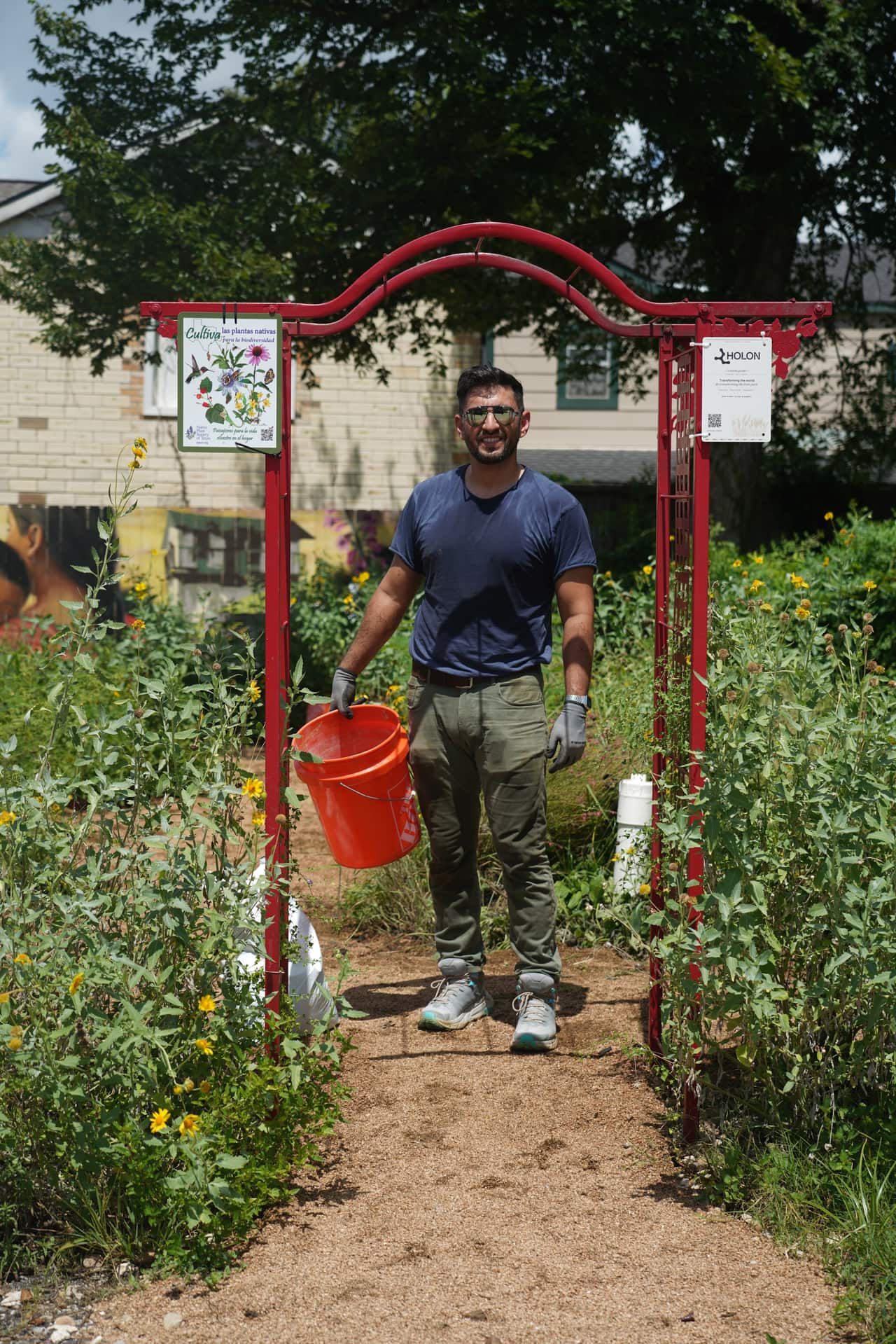 A neighbor standing under a red garden arch holding an orange bucket, garden all around