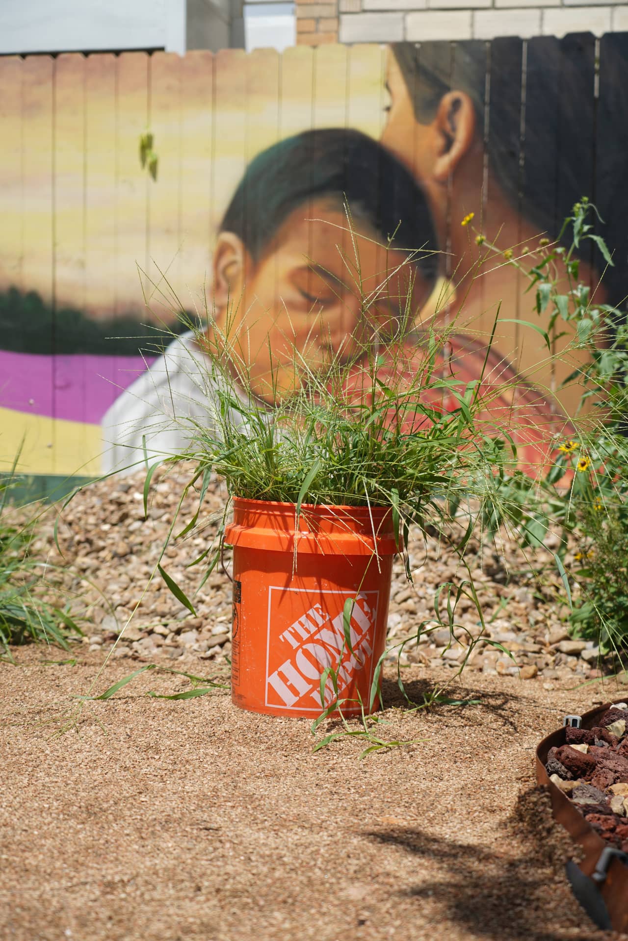 An orange work bucket filled with pulled grass on a gravel path in front of the mural